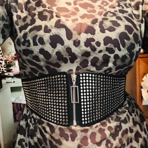 Brett midjebälte med nitar! - Snyggt brett elastiskt midjebälte i goth/rock-stil med dragkedja och nitar. Mått: B: 15-10 cm. L: 70-100 cm. Nytt!