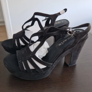 Tamaris sandaletter - Svarta sandaler från Tamaris med hög klack och öppen tå. Skorna har en elegant design med remmar och ett spänne runt ankeln för en säker passform. Perfekta för att ge en stilfull touch till din outfit.