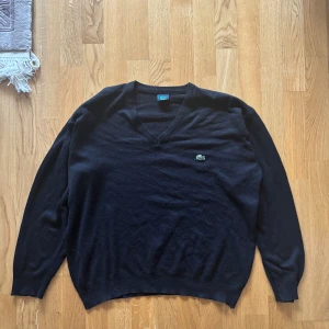 Svart tröja från Lacoste - Säljer en stilren svart tröja från Lacoste med v-ringning och det klassiska krokodilmärket på bröstet. Tröjan har långa ärmar och är perfekt för en avslappnad men ändå elegant look. Passar bra till både jeans och chinos.