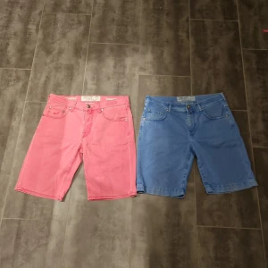 Färgglada shorts från Jacob Cohen - Säljer två par snygga shorts från Jacob Cohen i färgerna lax rosa och blå. De är tillverkade i Italien och har en klassisk design med fem fickor och knappgylf. Perfekta för sommaren när det är för varmt för jeans.