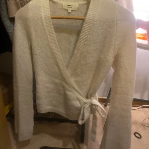 Vit omlottkofta från Gina Tricot - Säljer en mysig vit omlottkofta från Gina Tricot i ullblandning. Koftan har långa ärmar och knytband i midjan för en justerbar passform. Perfekt för kyliga dagar!