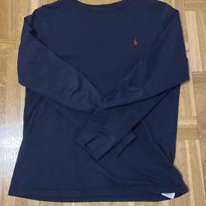 Mörkblå långärmad tröja från Polo Ralph Lauren - Säljer en stilren mörkblå långärmad tröja från Polo Ralph Lauren med det klassiska röda logotypbroderiet på bröstet. Perfekt för en avslappnad och snygg look. Tröjan är tillverkad i ett mjukt och bekvämt material.