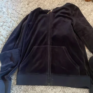 Svart velour hoodie från Juicy Couture - Säljer en svart velour hoodie från Juicy Couture med en stor broderad logga på ryggen. Tröjan har dragkedja framtill och långa ärmar. Perfekt för en avslappnad och stilren look.
