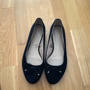 Svarta mocka ballerinaskor med rosett - Säljer ett par eleganta svarta ballerinaskor i mocka med en söt rosett på tån. Skorna har en rund tå och är perfekta för en stilren look.