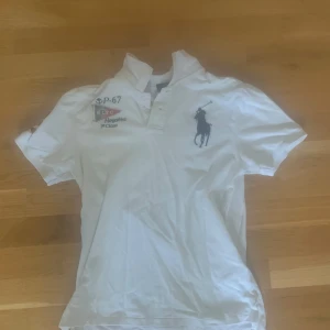 Ralph lauren tröja - Säljer en klassisk vit pikétröja från Ralph Lauren med korta ärmar och en stilren krage. Tröjan har ett stort tryck av en polospelare på framsidan och flera mindre tryck på ärmen och bröstet. Perfekt för en avslappnad och stilren look. Möts upp i Stockholm 