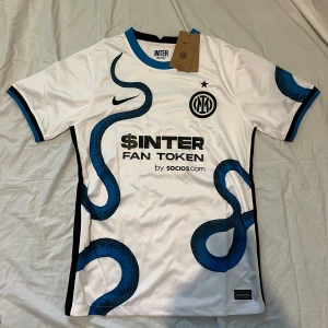 Vit och blå Inter fotbollströja - Säljer en vit Inter fotbollströja från Nike med blått ormmönster. Tröjan har korta ärmar med blå och svarta detaljer. Den har Inter-loggan och sponsortryck på framsidan samt Lenovo-tryck på baksidan. Perfekt för fans av laget!