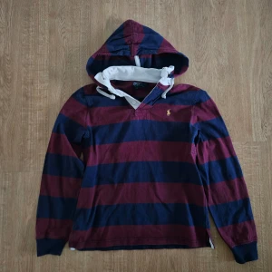Randig hoodie från Polo Ralph Lauren - Säljer en snygg randig hoodie från Polo Ralph Lauren i marinblått och vinrött. Tröjan har en klassisk design med långa ärmar och en praktisk huva. Den ikoniska loggan är broderad i gult på bröstet. Perfekt för en avslappnad stil!