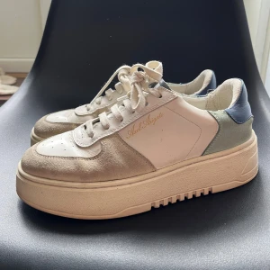 Sneakers från Axel Arigato - Snygga sneakers från Axel Arigato med en stilren design. Skorna har en beige och vit färgkombination med blå detaljer. Tillverkade i skinn och mocka med snörning och en rund tå. Perfekta för en trendig look.