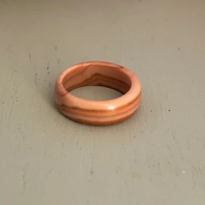 Ring med mönster - Säljer en unik ring i träfärgad design med naturliga ådringar. Ringen har en slät yta och en varm, jordnära ton som ger en naturlig känsla. Perfekt för dig som gillar en enkel men stilfull accessoar. Tror den är i glas men är osäker på materialet. 