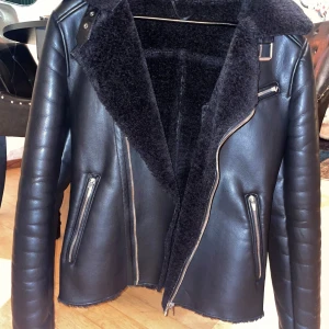 Svart skinnjacka från Zara Man - Snygg svart skinnjacka från Zara Man med mjukt foder och dragkedjor. Jackan har en cool bikerstil med en asymmetrisk dragkedja och flera fickor. Perfekt för en stilren look.