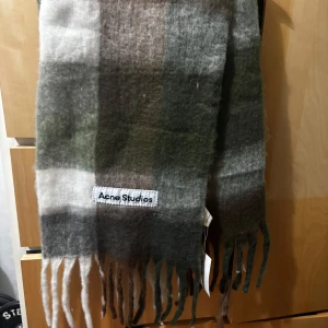 Acne Studios halsduk - Snygg och stilren rutig halsduk från Acne Studios i jordnära toner. Den har fransar i ändarna och en mjuk känsla som passar perfekt för kyliga dagar. Märkeslappen är tydligt synlig och ger en exklusiv touch.