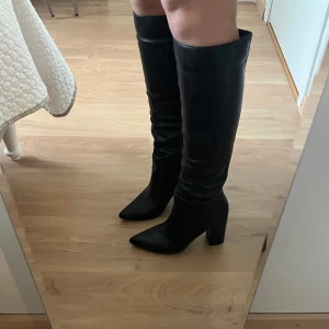 Svarta höga stövlar med klack  - Snygga svarta stövlar i skinnimitation med spetsig tå och bred hög klack. Stövlarna går upp till knäet och har en stilren, minimalistisk look som passar perfekt till kjol eller klänning. Klassisk modell med platt sula framtill och markerad klack. 