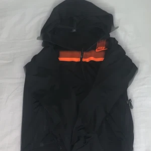 Nike svart vindjacka med orange detalj - Nike vindjacka i svart med en snygg orange detalj och logga på bröstet. Jackan har huva, dragkedja framtill och praktiska fickor med dragkedja. Perfekt för dig som gillar sportig stil och vill ha något lätt och funktionellt.