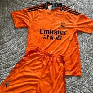Real Madrid Mbappé orange match kit - Snygg orange Real Madrid fotbolls kit med Mbappé och nummer 9 på ryggen. Tröjan har svarta detaljer, Adidas klassiska ränder på axlarna och klubbmärke på bröstet. Tillverkad i lätt och ventilerande material, perfekt för träning eller match.
