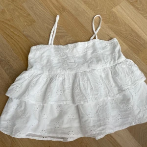 Vit volangtopp från Gina Tricot - Supersöt vit linnetopp från Gina Tricot med tunna axelband och broderade blommor. Toppen har volanger i flera lager och är perfekt för varma dagar. Lätt och luftig känsla, passar dig som gillar romantisk och somrig stil.