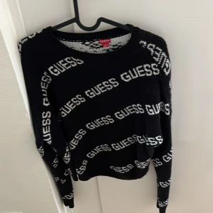 Svart stickad tröja från Guess - Säljer en svart stickad tröja från Guess med vit logotyptext över hela tröjan. Tröjan har rund halsringning och långa ärmar. Perfekt för dig som gillar tydliga loggor och en trendig look.