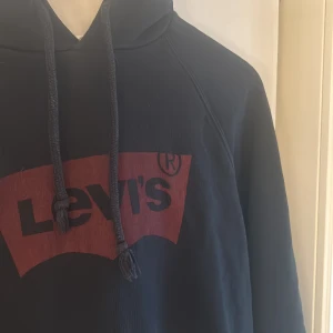 Svart hoodie från Levi's med logga - Svart hoodie från Levi's med stor röd logga framtill. Klassisk modell med huva och dragsnören, samt känguruficka. Mjuk insida och långa ärmar, perfekt för chill dagar. Passar dig som gillar streetwear och enkel stil.