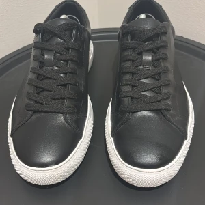 Svarta Hugo Boss skor med vit sula - Stilrena svarta sneakers med vit sula. Skorna har klassisk snörning och är tillverkade i ett skinnliknande material. Perfekta för en clean och enkel look. Hugo Boss skor. Skorna är inte stora i storleken.