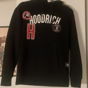 Svart hoodie från Hoodrich med tryck - Säljer en svart hoodie från Hoodrich med vita och röda tryck och patchar framtill. Tröjan har huva med dragsko och en stor ficka på magen. Perfekt för dig som gillar streetwear och vill ha något bekvämt och snyggt.