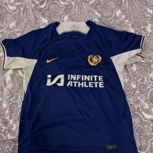 Chelsea FC blå fotbollströja från Nike - Säljer en blå Chelsea FC fotbollströja från Nike med klubbmärke på bröstet och Infinite Athlete som sponsortryck. Tröjan har korta ärmar med vita och guldiga detaljer samt rund hals. Perfekt för fotbollsfans!