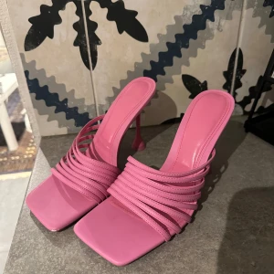 Rosa klackade sandaler med remmar - Snygga rosa sandaler med hög klack och flera tunna remmar över foten. Skorna har fyrkantig tå och öppen häl, vilket ger en trendig vibe. Tillverkade i syntetmaterial med glansig finish. Perfekta för dig som vill sticka ut med färg och stil. Klackhöjd 10cm