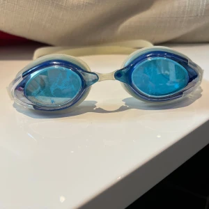 Blå simglasögon från Malmsten - Snygga simglasögon från Malmsten med blåtonade linser och vit transparent ram. Justerbart silikonband med svart logga baktill. Perfekta för simträning och tävling, ger bra sikt under vattnet och sitter skönt på huvudet.