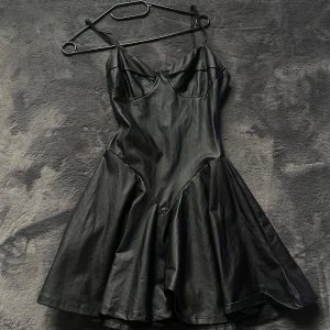 Svart kort klänning i skinnimitation - Snygg svart kort klänning i skinnimitation med smala axelband och markerad byst. Klänningen har en utställd kjol och figurnära passform upptill, vilket ger en cool och edgy look. Perfekt för dig som vill sticka ut med en trendig stil.