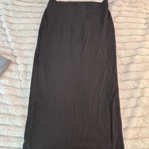 Svart ribbad långkjol Gina Tricot - Svart ribbad långkjol från Gina Tricot i storlek S. Kjolen har en rak och enkel passform med stretchigt material som sitter snyggt på kroppen. Perfekt till sneakers eller boots för en chill och stilren look. Kjolen går ner till lite över hälen.