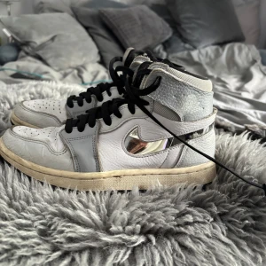 Nike Air Jordan 1 High Silver/White - Unika Nike Air Jordan 1 High med vit och ljusgrå ovandel i skinn och mesh, svarta snören och spegelblankt silver-Swoosh. Klassisk siluett med högt skaft och perforerad tå. Riktigt snygga detaljer.