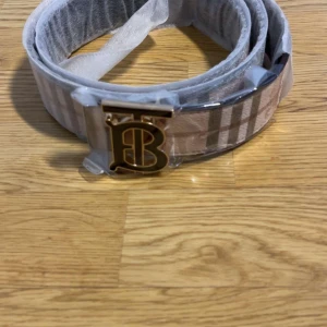 Beige bälte från Burberry med guldspänne - Säljer ett beige bälte från Burberry med klassiskt rutigt mönster och ett stort guldigt 'TB'-spänne. Bältet levereras med originalpåse, ask och certifikat. Perfekt accessoar för att lyfta din outfit.