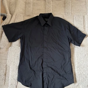 Svart kortärmad skjorta från Jobeline - Säljer en svart kortärmad skjorta från Jobeline med klassisk krage. Skjortan har en stilren och enkel design, perfekt för dig som gillar en clean look.