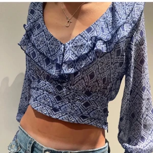 Blå mönstrad croppad blus med volang - Säljer en blå croppad blus med vitt mönster och volangdetalj längs den v-ringade halsen. Blusen har långa ärmar och är köpt ifrån Zara. Använd bara en gång, alltså nyskick! 