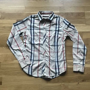 Burberry skjorta  - Snygg rutig skjorta från Burberry London i storlek S. Skjortan är beige med klassiskt rutmönster i blått och rött, har långa ärmar och knäppning framtill. Tillverkad i mjuk bomull och passar perfekt för en stilren look. Bara att skriva vid frågor :)