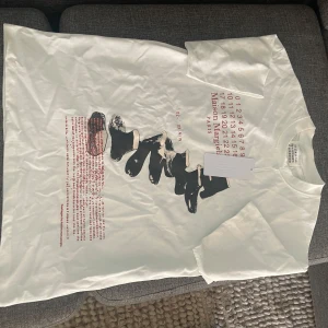 Maison Margiela t-shirt - Helt ny vit t-shirt från Maison Margiela med unikt svart och rött grafiskt tryck framtill samt ikoniska sifferdetaljer. Klassisk passform med rund hals och korta ärmar. Tillverkad i mjuk bomull för en clean och modern vibe. Beställde hem den från best secret men storleken passade inte. Nypris 2800kr
