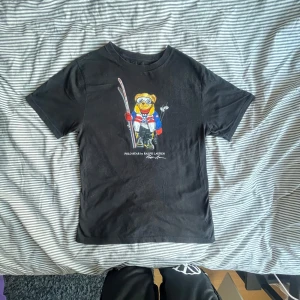 Svart Polo Bear t-shirt från Ralph Lauren - Svart t-shirt från Ralph Lauren med ikoniska Polo Bear-trycket framtill. Björnen har skidutrustning och färgglada kläder i rött, blått och vitt. Klassisk rund hals och korta ärmar. Skön bomullskvalitet, perfekt för dig som gillar streetstyle och coola prints.