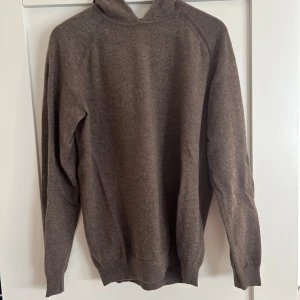 Brun hoodie i ullblandning - Mysig brun hoodie med lång ärm och huva. Tröjan är tillverkad i mjuk cashmere och har en enkel, stilren design utan tryck eller detaljer. Perfekt för kyliga dagar när du vill ha något bekvämt och snyggt.
