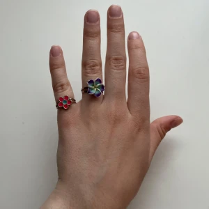 Två blommiga färgglada ringar - Säljer två söta ringar med blommotiv. Den ena ringen har en rosa blomma med turkos mitt, och den andra har en större blomma i lila, blått och vitt. Båda ringarna har justerbara silvriga band och är perfekta för att ge din stil en lekfull touch. 20 kr för båda, säljs ej separat!