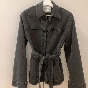 Svart jeansjacka från Carin Wester - Snygg svart eller mörkgrå jeansjacka från Carin Wester med krage, stora fickor framtill och knäppning med knappar. Jackan har ett bälte i midjan för en mer definierad siluett och långa ärmar. Nypris straks under 800kr!!!