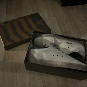 Vita sneakers med grå stjärndetalj - Säljer ett par vita sneakers från Golden Goose med grå stjärna på sidan och grå sula. Skorna har snörning och klassisk låg modell. Perfekta för dig som gillar stilrena och trendiga skor.