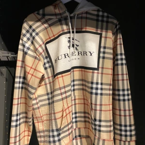 Rutig hoodie från Burberry - Säljer en klassisk beige hoodie från Burberry med det ikoniska rutmönstret i blått, rött och svart. Stor logga framtill och baktill, vit dragsko och känguruficka. Perfekt för dig som gillar streetwear med lyxig känsla.