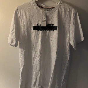 Vit Lacoste t-shirt med tryck - Vit t-shirt från Lacoste i slim fit-modell med svart tryck över bröstet. Klassisk rund halsringning och korta ärmar. Tillverkad i stretchig bomull för extra komfort. Perfekt för en clean och sportig look.