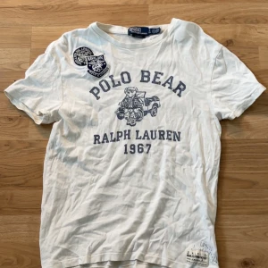Vit Polo Bear t-shirt från Ralph Lauren - Vit t-shirt från Polo Ralph Lauren med klassisk Polo Bear-tryck och texten 'Ralph Lauren 1967' framtill. T-shirten har korta ärmar, rund hals och coola patchdetaljer på axeln. Tillverkad i mjuk bomull för en skön känsla.
