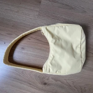 Ljusgul axelväska  - Snygg ljusgul axelväska med enkel och stilren design. Väskan har en mjuk, vadderad känsla och stängs med dragkedja upptill. Perfekt storlek för det viktigaste och lätt att slänga över axeln till vardags.
