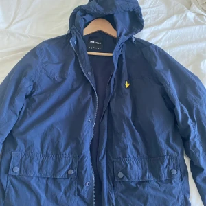 Blå vindjacka från Lyle & Scott - Säljer en blå vindjacka från Lyle & Scott med huva och dragkedja. Jackan har två framfickor med knappar och den klassiska gula loggan på bröstet. Perfekt för blåsiga dagar och enkel att matcha med olika outfits. Bra skick förutom ett litet hål i baksidan av jackan.
