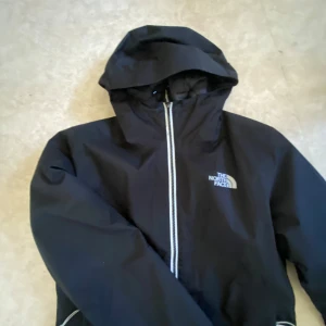 Svart vindjacka från The North Face - Säljer en svart vindjacka från The North Face med huva och hel dragkedja framtill. Jackan har logga på bröstet och ryggen, samt justerbar dragsko vid huvan. Perfekt för blåsiga dagar och enkel att matcha.
