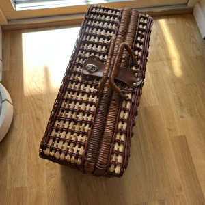 Brun och beige picknickkorg i rotting - Säljer en klassisk picknickkorg i flätad rotting med handtag och läderremmar. Korgen är brun och beige och har plats för tallrikar och muggar, perfekt för picknick. Insidan är klädd i blåvitrutigt tyg och har fack för porslin och bestick.