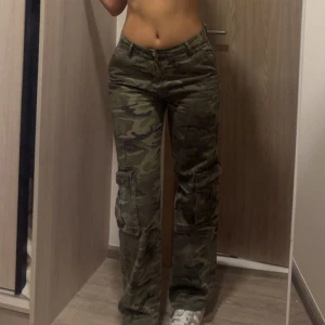 Cargobyxor i camouflage - Säljer ett par gröna cargobyxor med camouflage-mönster. Byxorna har flera fickor på sidorna och en rak, loose passform. 