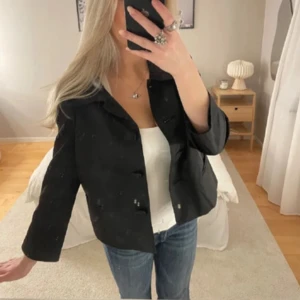 Svart kappa med stora knappar - Snygg svart kavaj med stora knappar framtill och krage. Jackan har en rak passform och trekvartsärmar, perfekt att slänga över en topp eller t-shirt. Passar till jeans eller kjol för en stilren look.