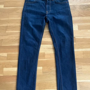 Mörkblå jeans från Nudie Jeans - Säljer ett par klassiska mörkblå jeans från Nudie Jeans med raka ben och snygga kontrastsömmar. Jeansen har fyra fickor och Nudie Jeans signaturbroderi på bakfickorna. Perfekta för dig som gillar stilrena och tidlösa jeans.