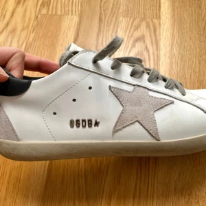 Vita sneakers med grå stjärndetalj från Golden Goose - Säljer ett par vita sneakers från Golden Goose med grå mockadetaljer och ikonisk stjärna på sidan. Skorna har svarta hälpartier, grå snörning och beige sula. Perfekta för en avslappnad och trendig stil. De flesta storlekar finns tillgängliga. Leverans sker inom 12–15 dagar.